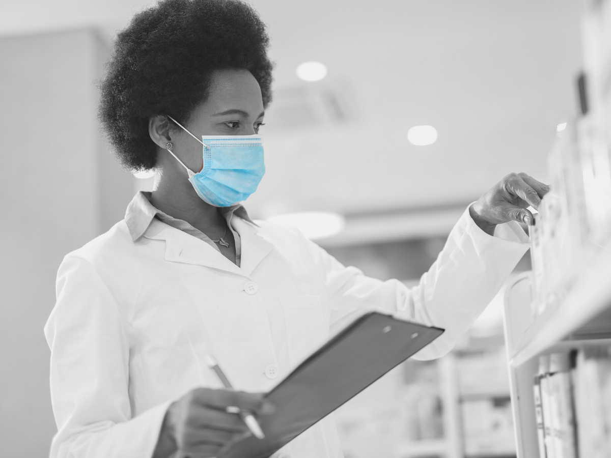 Image of a person wearing a lab coat, wearing a medical mask over their nose and mouth, while looking at items on a shelf and holding a clipboard.